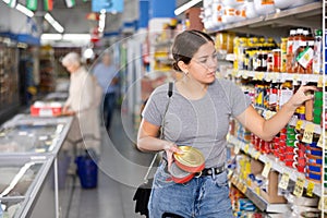 Young woman purchaser choosing tinned fish in big supermarket