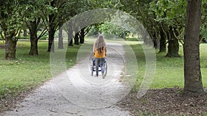 Young woman pushing a wheelchair through the middle of a tree-lined driveway in a park, rear view