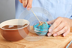 Young woman putting cake pop into blue sprinkles on board, closeup