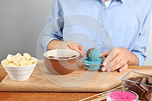 Young woman putting cake pop into blue sprinkles on wooden board