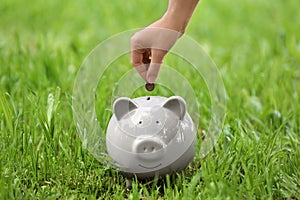 Young woman putting coin into piggy bank on grass outdoors, closeup