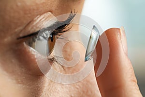 Young woman putting contact lens in her eye on blurred background, closeup