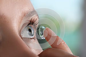 Young woman putting contact lens in her eye on blurred background, closeup