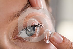 Young woman putting contact lens in her eye on blurred background, closeup