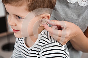 Young woman putting hearing aid in little son`s ear indoors