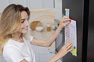 Young woman putting to do list on refrigerator in kitchen