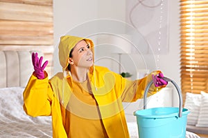 Young woman in raincoat collecting leaking water from ceiling at home. Time to call roof repair service