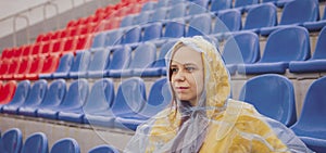 Young woman in raincoat sitting on stadium bleachers alone in rainy weather. Female spectator in wet waterproof coat at