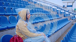 Young woman in raincoat sitting on stadium bleachers alone in rainy weather. Female spectator in wet waterproof coat at