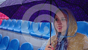 Young woman in raincoat with umbrella sitting on stadium bleachers alone in rain. Female spectator in transparent