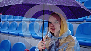 Young woman in raincoat with umbrella sitting on stadium bleachers alone in rain. Female spectator in transparent