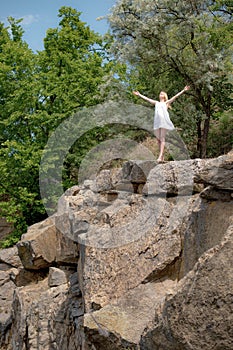 Young woman with raised hands sky on the edge of the cliff in summer day. Girl wearing short white dress.
