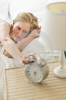 Young Woman Reaching For Alarm Clock While Lying On Bed