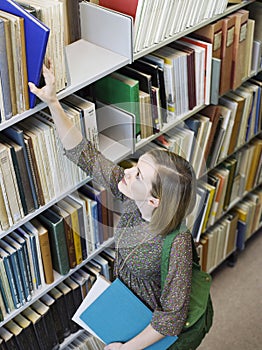 Young Woman Reaching For Book From Library Shelf