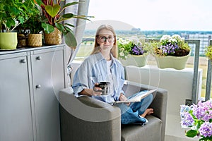 Young woman reading book drinking coffee sitting at home in armchair