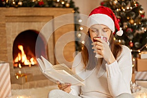 Young woman reading book and drinking hot beverage, relaxes in front of a blazing fire alongside Christmas tree and gifts, lady in