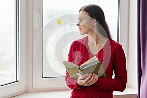 Young woman in reading book, girl stands near window with yellow fallen leaf