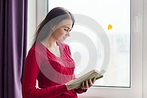 Young woman in reading book, girl stands near window with yellow fallen leaf