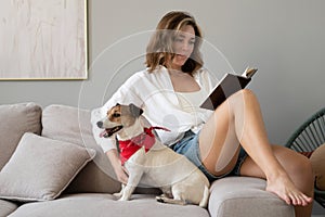 Young woman reading book and her cute dog on sofa at home. Lovely pet.