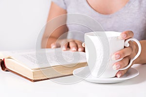 Young woman reading book and holding cup of coffee at home desk