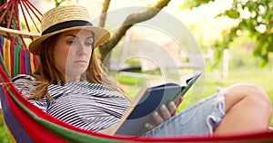 Young woman reading book while lying in comfortable hammock at green garden