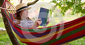 Young woman reading book while lying in comfortable hammock at green garden