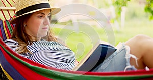 Young woman reading book while lying in comfortable hammock at green garden