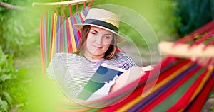 Young woman reading book while lying in comfortable hammock at green garden