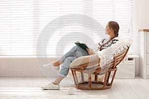 Young woman reading book in papasan chair near window at home