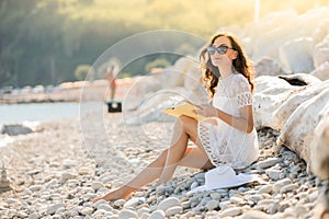 Young woman is reading a book on the pebble beach, with big white hat and dress