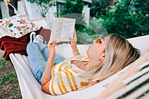 Young woman reading book, relaxing in hammock outdoors, in the backyard garden