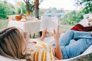 Young woman reading book, relaxing in hammock outdoors, in the backyard garden