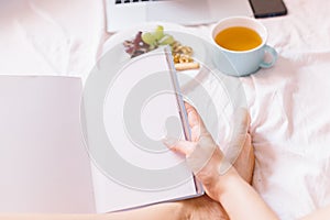 Young woman reading a book. Tea at home in her bed, checking her laptop, reading a book and having breakfast