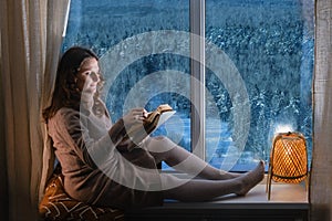 Young woman reading a book by the window in the light of the evening lamp. Woman sitting on the windowsill by the window with snow