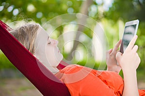 Young woman reading on electronic tablet reader relaxing in hammock under palm trees