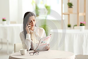 Young woman reading newspaper while talking on mobile phone in cafe