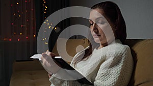 Young woman reads book sitting on sofa in room in evening