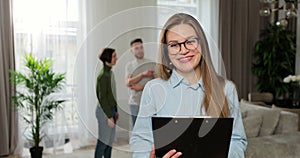 Young woman realtor looking at camera standing with papers while couple talking watching interior in background. Buying