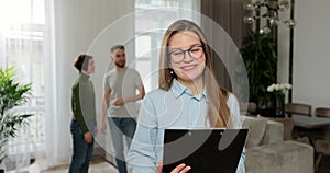 Young woman realtor looking at camera standing with papers while couple talking watching interior in background. Buying