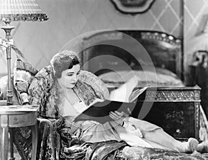 Young woman reclining in an armchair and reading a book in her bed room