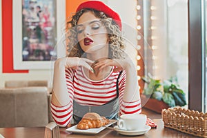 Young woman in red beret sitting in cafe, drinking coffee and eating french croissant