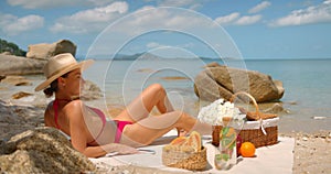 Young woman in red bikini and a hat sunbathing on beach lying on white towel overlooking the ocean