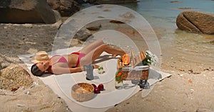 Young woman in red bikini sunbathing on isolated beach lying on white towel with picnic basket on it