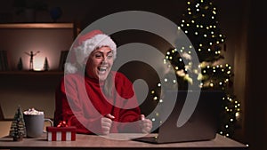 Young woman in red christmas santa hat very happy and rejoices sitting in front of laptop at the table . Christmas sales