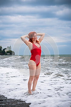A young woman in a red closed swimsuit. Stormy sea with big waves, Heavy thunderstorms on the horizon. Sunset. Rocky beach.