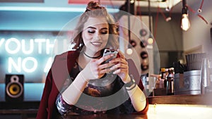 young woman in a red coat sitting alone by the bar, smiling and surfing in her phone. Fashionable outlook, amazing