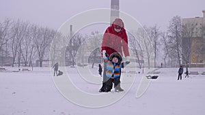 Young woman in red down jacket helps her baby boy to walk under the snow to the camera on the winter city park