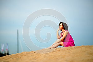 Young woman in a red dress ans curly hair sit on the sand