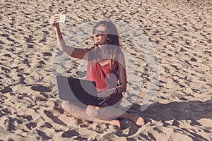 Young woman in red dress with computer and smartphone on the beach. Freelance and downshifting concept.