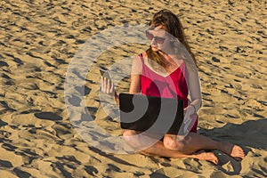 Young woman in red dress with computer and smartphone on the beach. Freelance and downshifting concept.